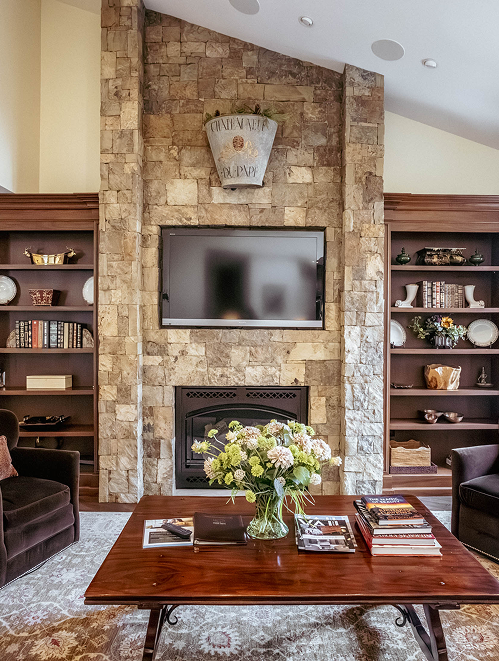 Cozy lodge interior featuring a stone fireplace, modern flat-screen TV, and stylish bookshelves, with a wooden coffee table adorned with floral arrangement and magazines, embodying the luxury and comfort of RamsHorn Lodge in Vail, Colorado.