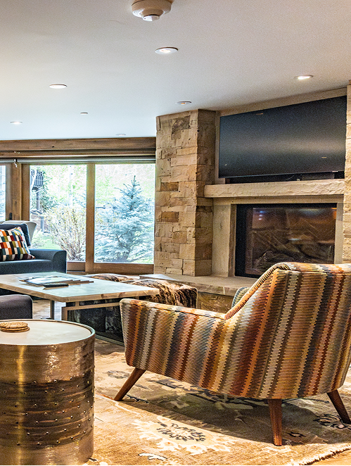 Cozy living area in Rams-Horn Lodge with modern mountain design, featuring a colorful armchair, stone fireplace, and large windows showcasing outdoor greenery.