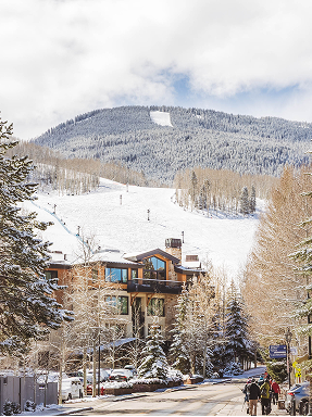 Scenic view of RamsHorn Lodge in Vail, Colorado, with snow-covered mountains and skiers in the background, highlighting the lodge's proximity to slopes and winter activities.